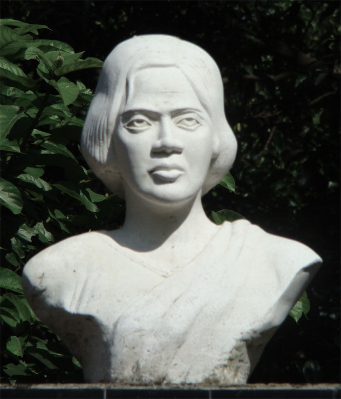 A white stone bust of Pritilata Waddedar with jaw-length hair and a draped sari, photographed against dark green foliage.