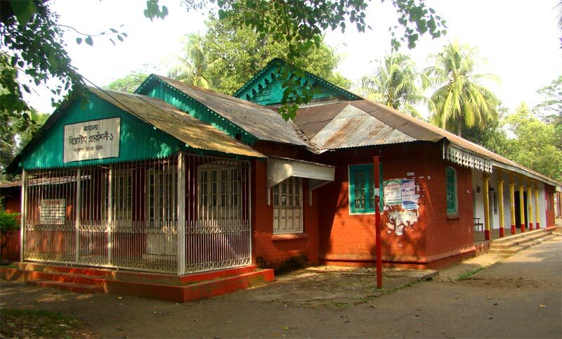 A low, single-storey colonial-era building with a green corrugated roof, red walls and a railed verandah, surrounded by trees.