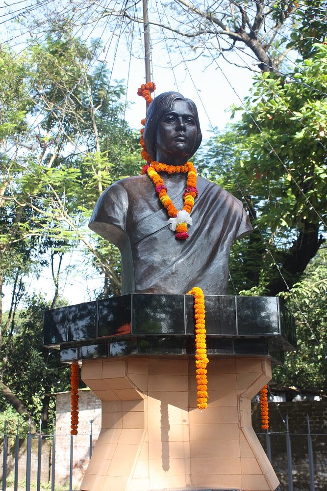 A dark bronze bust of Pritilata Waddedar on a stepped plinth hung with marigold garlands, standing outdoors among trees with commemorative streamers overhead.