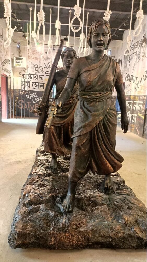 A bronze-toned sculpture of two women in saris striding forward in a museum gallery, with the painted names of revolutionaries covering the walls behind.